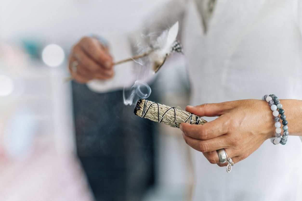 Woman’s hand holding bundle of dried sage herb, performing smudging ritual, cleansing negative energy and purifying living space with smoke